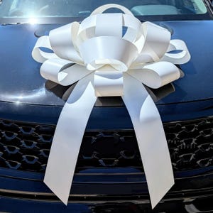 May include: A large, white decorative bow, possibly for a car, sits on the bonnet of a dark-coloured vehicle. The bow is made of wide, satin-like ribbons, with long, flowing tails. The bow's design is intricate and eye-catching.