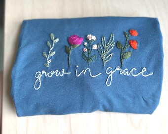 Puede incluir: Un artículo de tela azul doblado con diseños florales bordados y las palabras "grow in grace" en escritura blanca. El bordado presenta una rosa rosa, pequeñas flores blancas y flores naranjas.