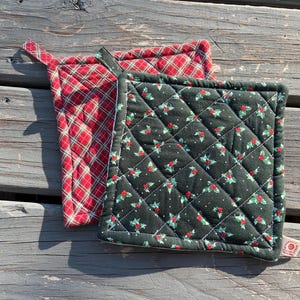 May include: Two quilted pot holders. One is red with a plaid pattern, and the other is dark green with a holly and berry design. Both have a square shape and a loop for hanging. The pot holders are on a weathered wooden surface.