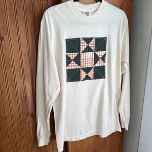 Puede incluir: Camiseta de manga larga color crema con un diseño de bloque de edredón cuadrado. El bloque de edredón presenta un borde verde oscuro, con patrones de cuadros y estrellas de color naranja y crema. La camiseta está colgada en una percha negra.