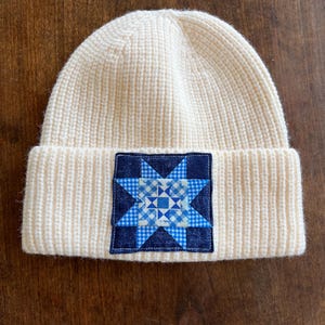 Puede incluir: Gorro de punto color crema con borde doblado. Un parche cuadrado en la parte delantera presenta un diseño de edredón de estrella azul y blanco. El gorro está hecho de punto acanalado.