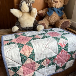 Peut inclure: Une courtepointe faite à la main avec un motif d'étoile en rose, vert et blanc, avec une bordure bleue. Deux jouets en peluche, un ours en peluche marron avec un nœud bleu et un lapin gris et blanc, sont posés au-dessus de la courtepointe.