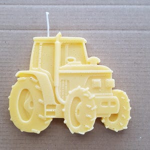 以下が含まれることがあります： 白い芯が付いた黄色のワックス製のトラクターのキャンドル。キャンドルは、フロントグリル、車輪、運転席が詳細に描かれています。