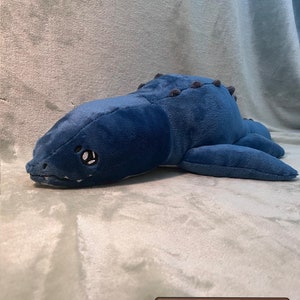 May include: A plush blue and gray dinosaur toy with a toothy grin and a spiky back. The toy is lying on a light gray fuzzy surface.  PDF Pattern.
