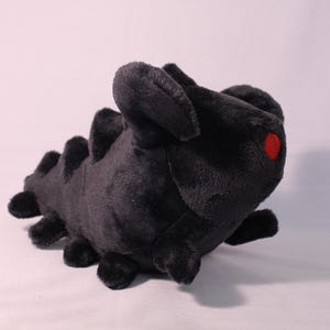 May include: A black plush toy shaped like a worm with a red eye. The toy has a segmented body and a textured surface.