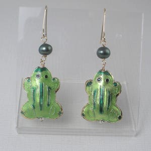 May include: A pair of green frog dangle earrings with black pearl accents. The frogs are made of enamel and have gold accents.