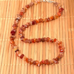 May include: A necklace composed of small, irregularly shaped orange and red gemstones. The necklace is strung on a silver chain and clasp. The gemstones are in shades of orange, red, and brown. The necklace is displayed on a bamboo mat.