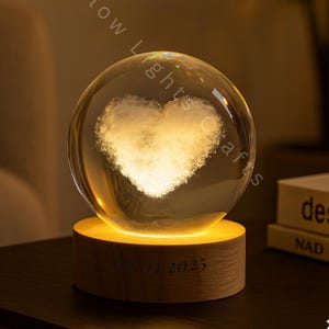 May include: A clear crystal ball with a heart-shaped design illuminated from within, resting on a wooden base. The base has the text "07.2023" engraved on it. The warm light creates a cozy ambiance. The product is from Glow Lights Crafts.