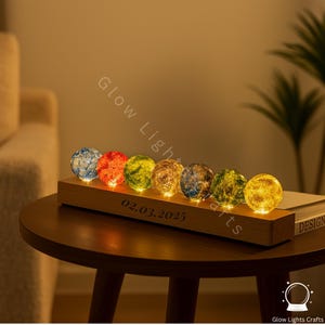 以下が含まれることがあります： 木製の台座に、6つのカラフルなガラス球が置かれ、内側から光を放っています。球体は青、赤、緑、黄、金の色合いで光っています。台座には「02.03.2025」の文字が刻まれています。装飾用のライトです。