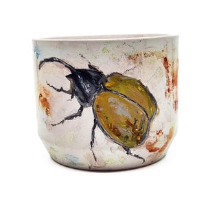 May include: A ceramic planter with a hand-painted beetle design. The beetle has a black head and legs with olive-green wings. The planter is off-white with brown and orange paint strokes.