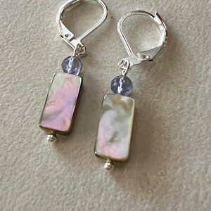 Può includere: Un paio di orecchini con chiusura a leva. Ogni orecchino presenta un pendente rettangolare in madreperla iridescente con sfumature rosa e oro, una piccola perla blu e una chiusura a leva color argento. Una piccola perla d'argento è in fondo a ogni pendente.