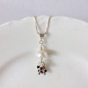 May include: A silver necklace with a pendant featuring a large, white, teardrop-shaped pearl. Below the pearl are clusters of small, round, black and red beads. The necklace is displayed on a white, scalloped plate.