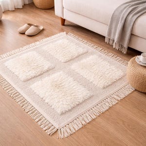Puede incluir: Alfombra rectangular color crema con cuatro acentos cuadrados y esponjosos y un borde con flecos. La alfombra está colocada sobre un suelo de madera, con un sofá y objetos decorativos en el fondo. Un elemento de decoración del hogar.
