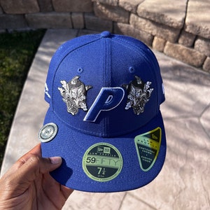 May include: A royal blue baseball cap with a white "P" logo and two black and white graphic designs. The cap has a black and gold "New Era" logo and a black and gold "59FIFTY" label with the size "7 1/4" printed on it. The cap also has a black and gold label that says "Low Profile" and "Contoured Crown Curved Visor".