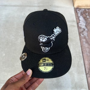 Op de afbeelding: Zwarte baseballpet met een wit geborduurd logo van een cartoonfiguur dat een honkbalknuppel vasthoudt. De pet heeft een zwarte klep en een gouden New Era 59FIFTY-logo met maat 7 1/4.