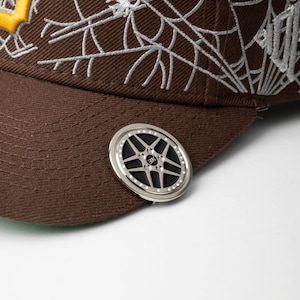 May include: A brown baseball cap with a white spiderweb design and a silver golf ball holder attached to the brim. The holder features a black rim and a silver wheel design.