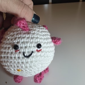 May include: White crocheted amigurumi toy with pink limbs and a smiling face. The toy has black eyes and a yellow button on its belly.