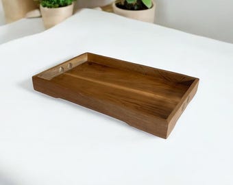 May include: A rectangular wooden tray with a natural wood grain finish. The tray has a raised edge and small circular cutouts on the corners. The tray is designed for serving or displaying items.