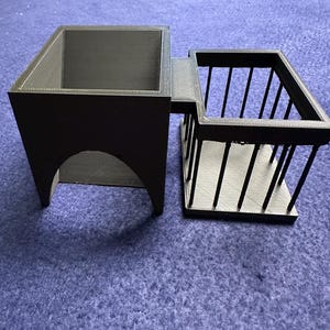 May include: A black 3D printed dice tower with a separate compartment for dice. The tower has a square base and a rectangular opening on the side. The compartment has a cage-like design with vertical bars.