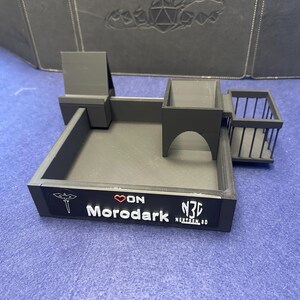 May include: Black 3D printed dice tray with a tower, a wall, and a balcony. The tray has a Morodark logo and the text "ON" on the front.