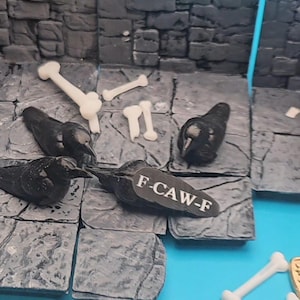 以下が含まれることがあります： 黒いカラスのフィギュア、骨型のオブジェクト、石壁の背景を特徴とするミニチュアジオラマシーン。カラスの1羽には「F-CAW-F」と書かれたタグが付いています。シーンは青い背景に設定されており、ファンタジーまたはテーブルトップゲームの美学を作り出しています。