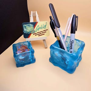 May include: Two blue resin ice cube shaped pencil holders. One holder contains dice, the other contains pens and a marker. The marker is a black Sharpie fine point marker.