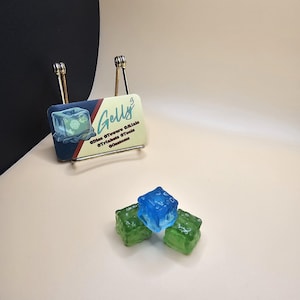 May include: Three dice, two green and one blue, with a frosted, translucent finish. The dice are sitting on a light beige surface. A business card for "Gelly's" is in the background.