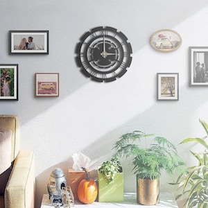 Può includere: Un orologio da parete nero e circolare con numeri romani e un design decorativo. L'orologio è montato su una parete bianca, circondato da quadri e oggetti decorativi, tra cui piante e una zucca.