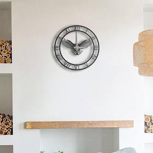 May include: A black metal wall clock with Roman numerals and an eagle design. The clock features a circular frame with the numbers, and the eagle is centered within the clock face. The clock is mounted on a white wall.