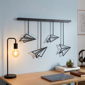 May include: A black metal wall art piece featuring five paper airplane designs suspended from a horizontal bar. The artwork is mounted on a light blue wall above a wooden desk. A black desk lamp with a round bulb is on the desk.