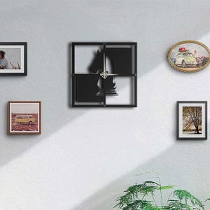May include: A black metal wall clock with a horse head design, surrounded by a square frame. The clock is mounted on a white wall, alongside framed pictures of various sizes and shapes. A green plant is visible at the bottom.