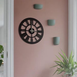 Può includere: Un orologio da parete nero e circolare con numeri romani e un design decorativo è montato su una parete rosa. Tre piccoli vasi da parete verde chiaro sono montati anche sulla parete. Una pianta verde si trova nell'angolo in basso a destra.