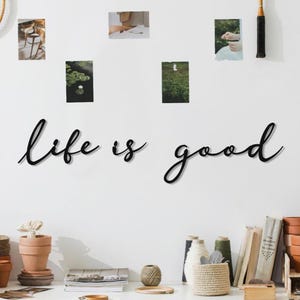 May include: Black cursive wall art that reads "life is good" is mounted on a white wall. The wall is decorated with small framed photos. Below the wall art is a white desk with books, pottery, and decorative items.