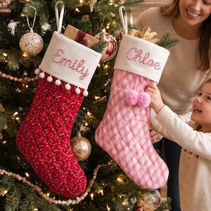 Può includere: Due calze natalizie trapuntate appese a un albero. La calza sinistra è rossa con un risvolto bianco ricamato con il nome Emily e bordino a pompon bianchi. La calza destra è a quadri rosa con risvolto bianco ricamato con il nome Chloe e due pompon rosa.