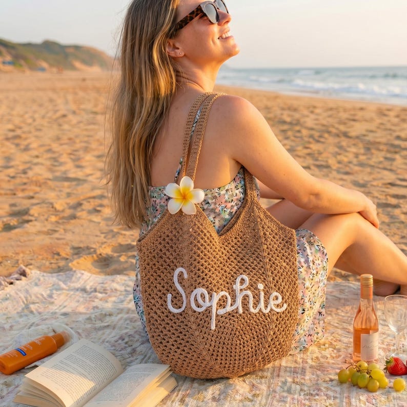Peut inclure: Un sac fourre-tout tiss&eacute; marron avec le nom "Sophie" brod&eacute; en &eacute;criture blanche. Le sac a deux anses et est orn&eacute; d'une fleur blanche et jaune. Le sac est sur une plage avec une femme portant des lunettes de soleil.