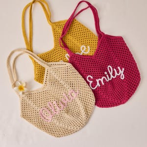 Peut inclure: Trois sacs fourre-tout en crochet jaune, fuchsia et beige. Chaque sac a une longue anse et un nom brod&eacute; sur le devant dans une couleur contrastante : "Olivia", "Emily" et "Love". Une fleur blanche et jaune est attach&eacute;e au sac beige.