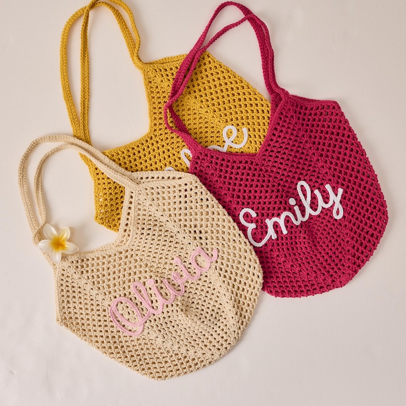 Peut inclure: Trois sacs fourre-tout en crochet jaune, fuchsia et beige. Chaque sac a une longue anse et un nom brod&eacute; sur le devant dans une couleur contrastante : "Olivia", "Emily" et "Love". Une fleur blanche et jaune est attach&eacute;e au sac beige.