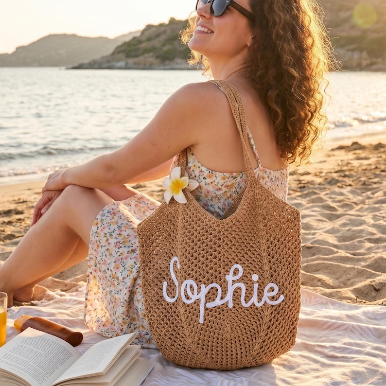 Peut inclure: Un sac fourre-tout en osier marron avec le nom "Sophie" brod&eacute; en blanc. Le sac a une bandouli&egrave;re et est orn&eacute; d'une fleur blanche. Le sac est sur une plage.