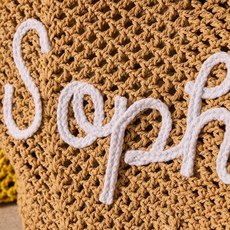 Peut inclure: Gros plan sur un tissu crochet&eacute; beige avec le nom "Soph" brod&eacute; en blanc. Les lettres sont faites d'un fil blanc &eacute;pais et sont cousues sur le tissage ouvert du tissu. L'arri&egrave;re-plan est l&eacute;g&egrave;rement flou.