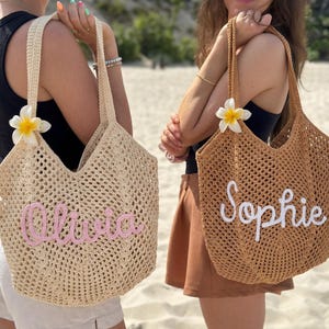Peut inclure: Deux sacs fourre-tout en tissu ajour&eacute;, un beige et un marron, chacun avec un nom brod&eacute; sur le devant. Le sac beige porte "Olivia" en rose, et le sac marron porte "Sophie" en blanc. Les deux sacs ont de longues anses et une fleur blanche et jaune.