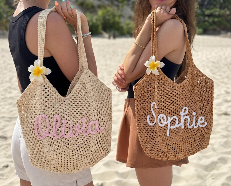 Peut inclure: Deux sacs fourre-tout en tissu ajour&eacute;, un beige et un marron, chacun avec un nom brod&eacute; sur le devant. Le sac beige porte "Olivia" en rose, et le sac marron porte "Sophie" en blanc. Les deux sacs ont de longues anses et une fleur blanche et jaune.