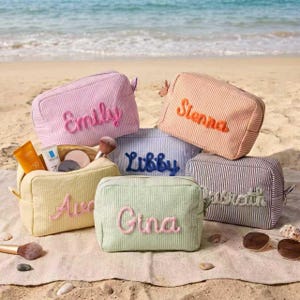 May include: A collection of five rectangular zippered pouches made of striped fabric in pastel colors including pink, peach, yellow, light blue, and lavender. Each pouch features a name embroidered in a contrasting color on the front surface.