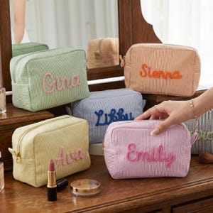 May include: A collection of striped cosmetic bags in various colors, each embroidered with a name. The bags are in shades of green, yellow, blue, orange, and pink. A gold zipper and pull complete the design. Makeup items are visible on the wooden surface.