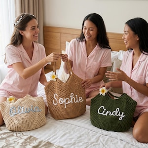 Peut inclure: Trois sacs fourre-tout tiss&eacute;s personnalis&eacute;s beiges, marron et verts, chacun avec un nom brod&eacute; en blanc : "Olivia", "Sophie" et "Cindy". Les sacs sont d&eacute;cor&eacute;s d'une fleur et sont tenus par trois personnes portant des pyjamas roses.