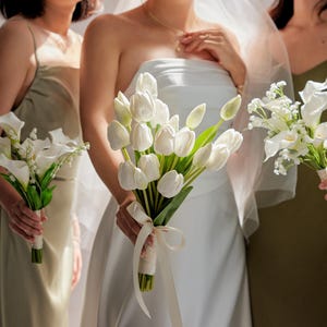 May include: A bride in a white strapless gown holds a bouquet of white tulips, with bridesmaids holding bouquets of white calla lilies and other white flowers. The bouquets are tied with ribbons. The women are standing in front of a window.