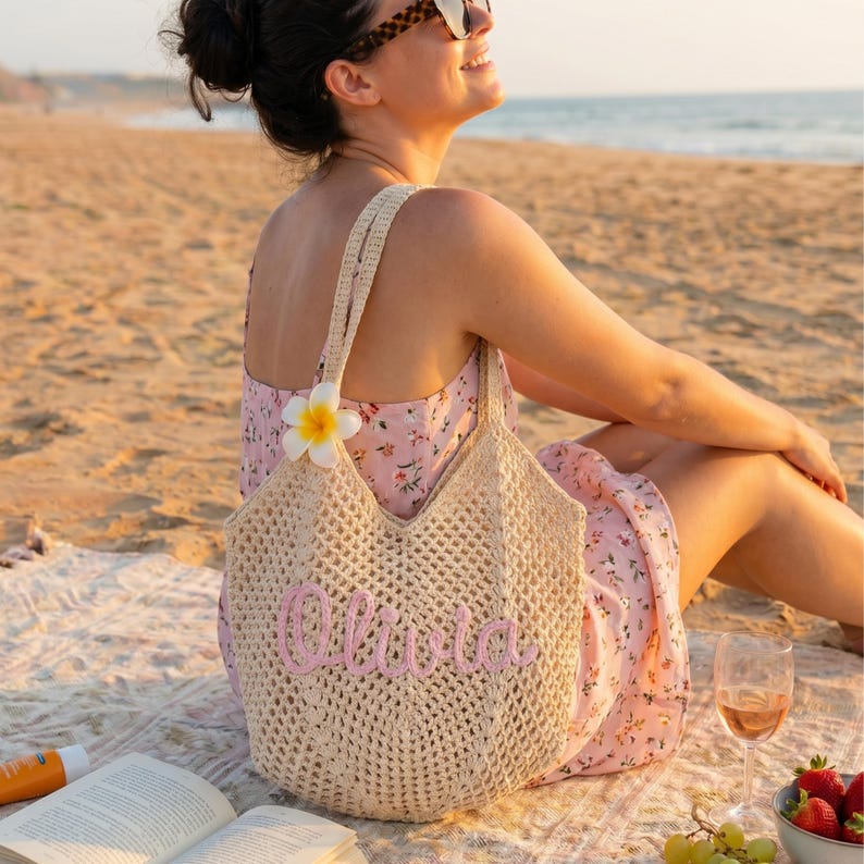 Peut inclure: Un sac fourre-tout en crochet beige avec le nom "Olivia" brod&eacute; en rose. Le sac a une ouverture en forme de V et de longues bretelles. Une fleur blanche et jaune est attach&eacute;e &agrave; la bretelle. Le sac est sur une couverture de plage.