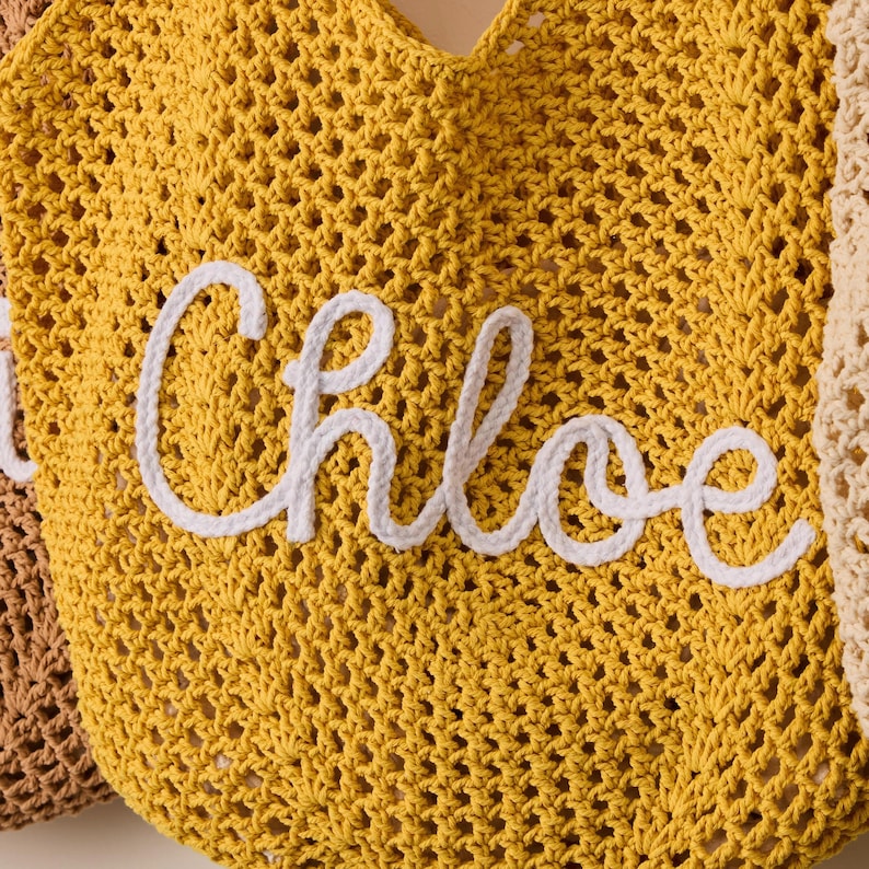 Peut inclure: Un sac fourre-tout en crochet jaune moutarde avec le nom "Chloe" en lettres de corde blanche. Le sac a un grand motif ajour&eacute; et le nom est centr&eacute; sur le devant. Le sac est de conception l&eacute;g&egrave;re et a&eacute;r&eacute;e.