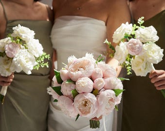 May include: A bride and bridesmaids hold bouquets of flowers. The bride's bouquet features large pink peonies, while the bridesmaids hold bouquets of white roses and pink accents. The women are wearing dresses in shades of white and olive green.