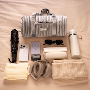 Peut inclure: Un sac de sport matelassé gris clair, un parapluie noir, une bouteille d'eau blanche, une coque de téléphone blanche, une paire de lunettes de soleil noires, un étui rectangulaire blanc, un casque audio supra-auriculaire gris, une écharpe en tricot beige et un téléphone blanc avec un écran noir.