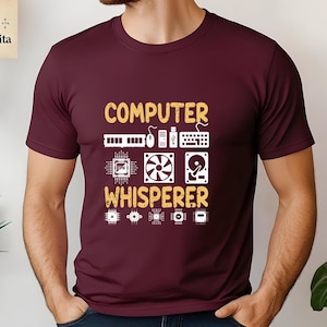 May include: Burgundy t-shirt with the text "COMPUTER WHISPERER" in yellow. The design includes a variety of computer hardware icons, such as a keyboard, mouse, hard drive, and circuit boards.
